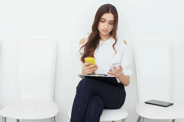 La fille est assise sur une chaise blanche parlant au téléphone et écrivant dans la tablette. Multitâche — Photo
