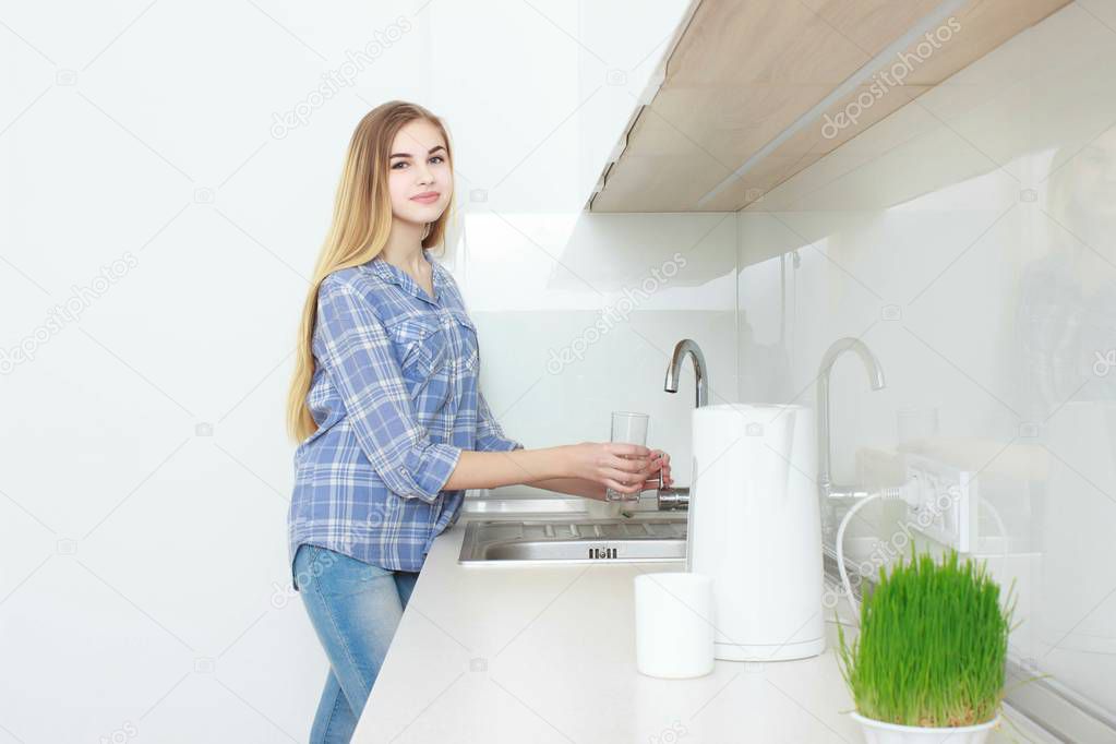 Young beautiful girl in a blue checkered shirt and jeans in the kitchen pouring the glass of water. Morning. home interior.