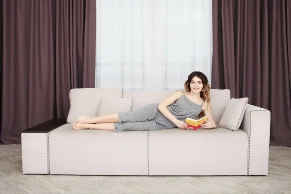 Mujer joven y bonita disfrutando leyendo un libro en casa tumbada en un sofá —  Fotos de Stock