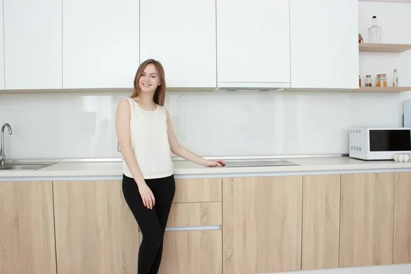 Красивая молодая женщина в белой футболке стоит на кухне и улыбается — стоковое фото