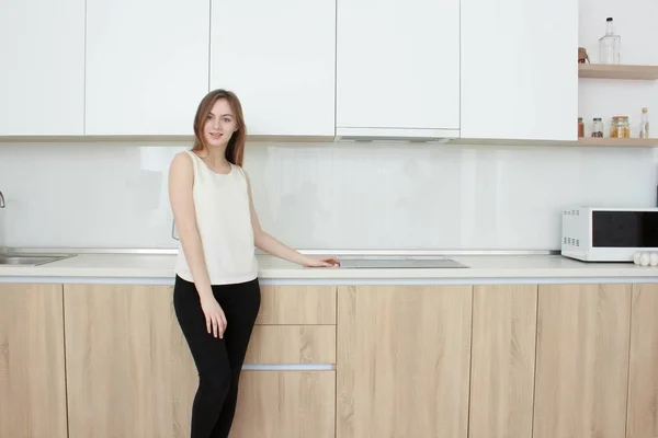 Красивая молодая женщина в белой футболке стоит на кухне и улыбается — стоковое фото