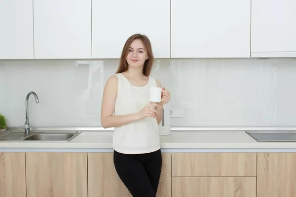 Молодая женщина в белой футболке, пьет кофе на кухне — стоковое фото