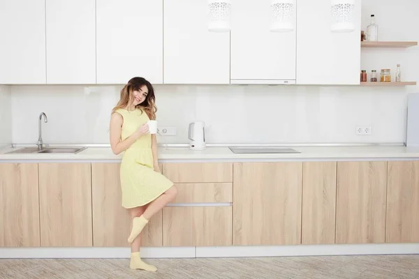 Ung kvinna i en gul klänning som dricker kaffe i köket. — Stockfoto