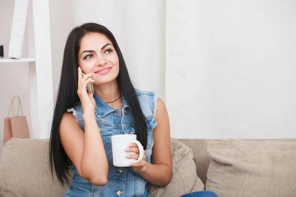 Piękna kobieta siedzi na kanapie, trzymając kubek gorącej herbaty i mówiąc telefon — Zdjęcie stockowe