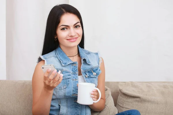 Jeune femme lisant des messages mobiles tout en tenant tasse de thé et assis sur le canapé — Photo