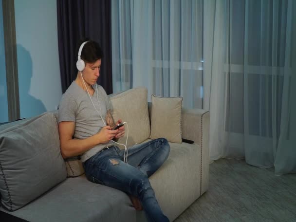 英俊的男人坐在沙发上放松，听听音乐带着耳机 — 图库视频影像