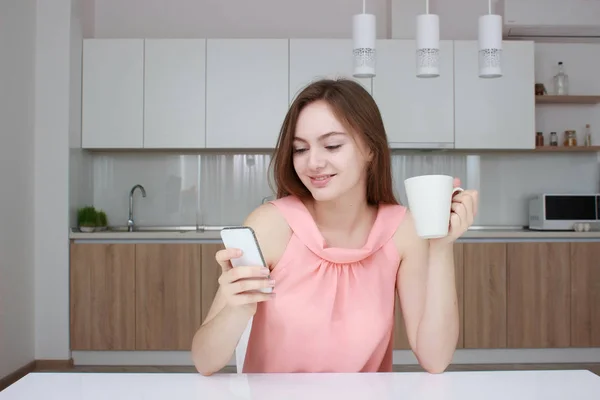 Beautiful girl in home using smartphone, drinking tea and smiling in kitchen — Stock Photo, Image