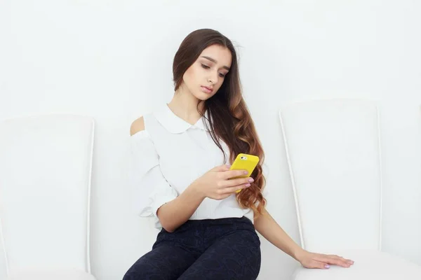 Улыбающаяся молодая деловая женщина сидит на стуле в современном офисе, пользуется смартфоном и пишет в блокноте — стоковое фото