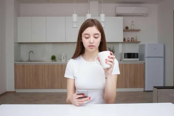 Young beautiful woman using cell phone and having a coffee in the kitchen — Stock Photo, Image