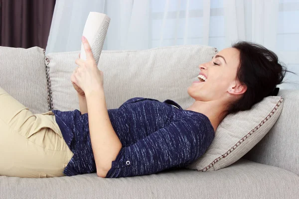 Belle brune souriante se relaxant sur le canapé et lisant un livre dans le salon — Photo