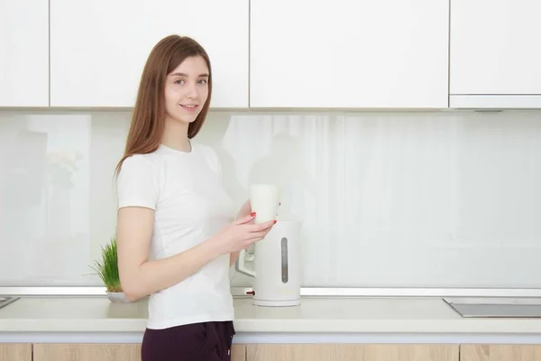 Красивая молодая женщина в белой футболке стоит на кухне и улыбается . — стоковое фото