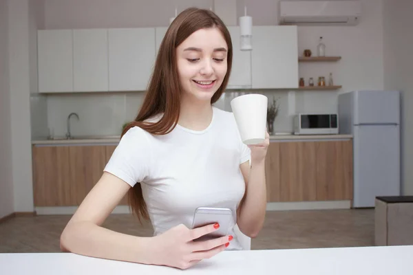 Young beautiful woman using cell phone and having a coffee in the kitchen