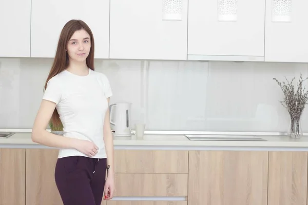 Mujer joven bonita en una camiseta blanca de pie en la cocina y sonriendo . — Foto de Stock