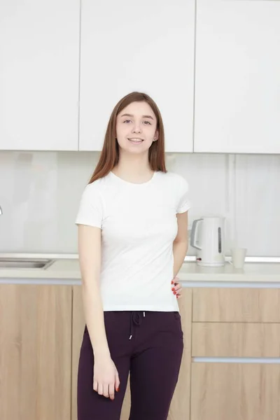 Досить молода жінка У білій сорочці, що стоїть на кухні і посміхається . — стокове фото
