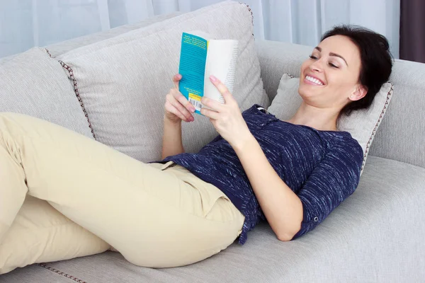Belle brune souriante se relaxant sur le canapé et lisant un livre dans le salon — Photo