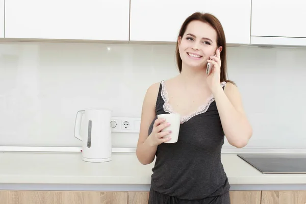 Brunette femme au foyer en blanc dans la cuisine parlant sur le téléphone mobile — Photo