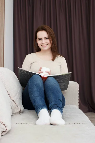 Ung kvinna hemma sitter på soffan avkopplande i sitt vardagsrum läsa bok — Stockfoto