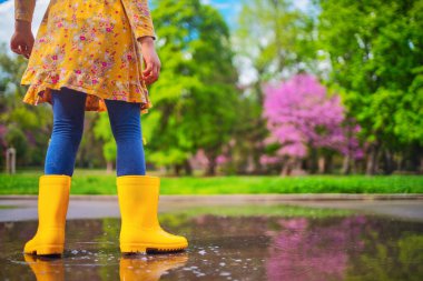Sarı lastik çizmeli mutlu kız çocuğu bahar yürüyüşünde su birikintisine atlıyor..