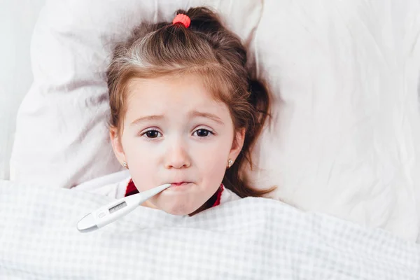 Beteg lány egy lázmérővel a szájában fekszik az ágyban. — Stock Fotó