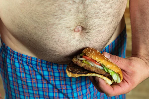 Un uomo che tiene un hamburger delizioso e succoso contro la sua pancia grassa. Anti fast food e tempo per un concetto di dieta . — Foto Stock