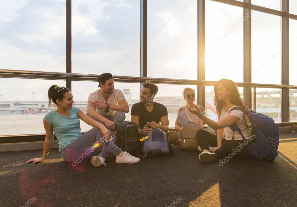 Young People Group Sitting On Floor In Airport Lounge Windows Waiting Departure Speaking Happy Smile Mix Race Friends