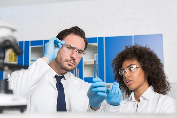 Dos científicos de laboratorio examinando líquido en tubo de ensayo, trabajadores de laboratorio de raza mixta estudian los resultados de la investigación química — Foto de Stock