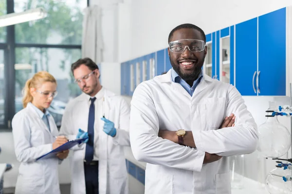 Cientista afro-americano sorridente feliz fica na frente de colegas em laboratório fazendo notas de experiência ou resultados de pesquisa — Fotografia de Stock