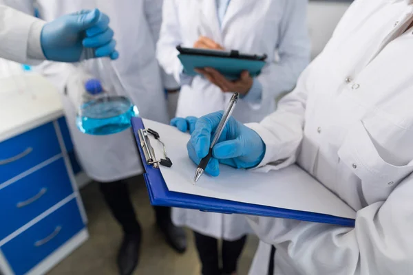 Primer plano del científico sosteniendo el frasco y el grupo de estudiantes tomando notas sobre el líquido en el laboratorio, equipo de investigadores escribiendo resultados del experimento — Foto de Stock