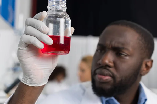 Científico afroamericano examina frasco con lubricante rojo trabajando en laboratorio moderno, investigador masculino haciendo experimento — Foto de Stock