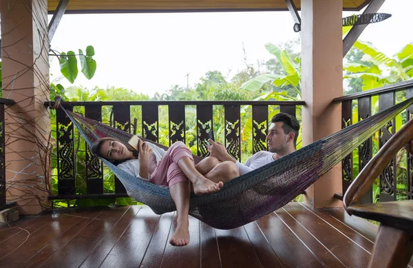 Jeune couple allongé dans l'hamac sur la terrasse hôtel tropical, homme et femme utilisant téléphone intelligent cellulaire bavardage vacances tropicales — Photo