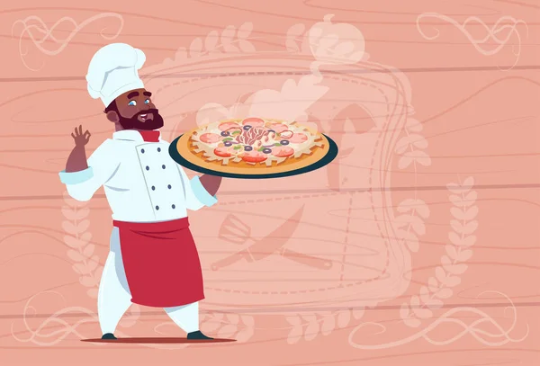 Chef afroamericano cocinero sosteniendo pizza sonriente jefe de dibujos animados en uniforme de restaurante blanco sobre fondo texturizado de madera — Vector de stock