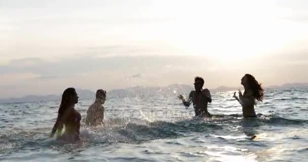 Grupo de amigos salpicando en el mar al atardecer, jóvenes divirtiéndose en el agua en la playa durante las vacaciones — Vídeo de stock