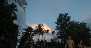 Gün batımı, mutlu erkek ve kadın deniz kenarında palmiye ağaçları konuşurken, sahilde insanlar yürüyüş grubu