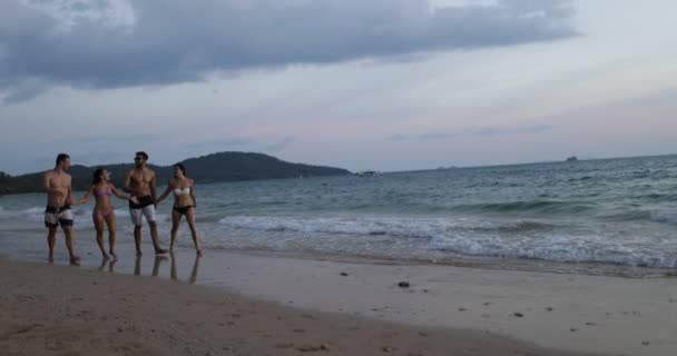 Zwei Paare, die bei Sonnenuntergang am Strand spazieren gehen und sich unterhalten, fröhliche Männer und Frauen, die gemeinsam auf Touristen am Meer zugehen — Stockvideo