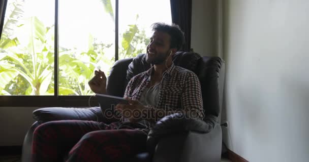 Młody człowiek z Tablet komputer słuchać o muzyki siedząc w fotelu w domu, wymieszać rasy facet noszenia słuchawek śpiewu — Wideo stockowe