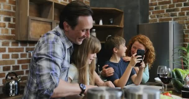 Familia junta en la cocina Padre e hija cortan verduras mientras el hijo y la madre usan el teléfono inteligente celular Riendo padres con dos hijos preparando comida — Vídeo de stock