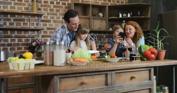 Familia juntos en la cocina Padre ayudando a su hija a cortar verduras mientras el hijo y la madre usan el teléfono inteligente celular Hablar con los padres con dos hijos Preparación de alimentos en casa — Vídeos de Stock