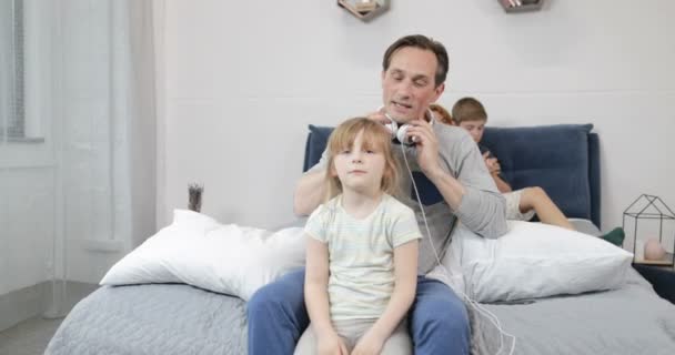 Padre poniendo nuevos auriculares en la pequeña hija escuchando música a través de auriculares mientras pasa tiempo en el dormitorio con la familia feliz — Vídeo de stock