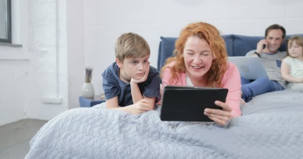 Familia pasar la mañana juntos en dormitorio madre e hijo ver vídeos riéndose más padre hablando por teléfono llamada abrazando hija — Vídeos de Stock