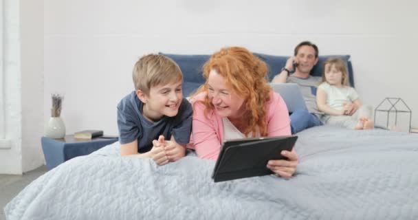 Мать и сын смеются с помощью планшетного компьютера смотреть смешные видео, лежащие на кровати в спальне семьи вместе — стоковое видео