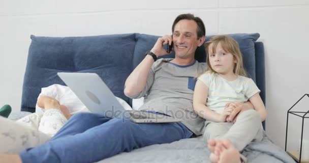 Отец обнимает маленькую дочь во время телефонного разговора счастливая улыбающаяся семья сидит вместе на кровати — стоковое видео