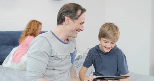 Happy Father And Son Use Tablet Computer Laugh Sitting On Bed In Bedroom Spending Time Together — Stock Video