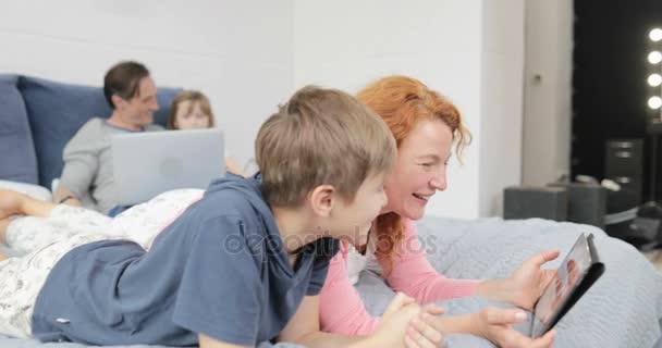 Мать с сыном смеются смотреть смешное видео на цифровом планшетном компьютере лежа на кровати, счастливая семья проводит время вместе дома — стоковое видео