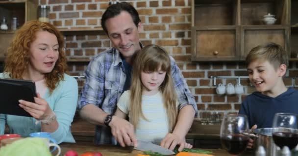 Szczęśliwa rodzina Kuchnia Gotowanie, ojciec i córka do krojenia warzyw, podczas gdy matka przeglądania przepis w Internet z cyfrowego tabletu — Wideo stockowe