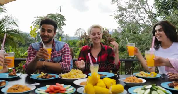 Gente tostando vasos de jugo sentados en la mesa en la terraza comiendo jóvenes amigos hablando al aire libre Comunicación — Vídeos de Stock