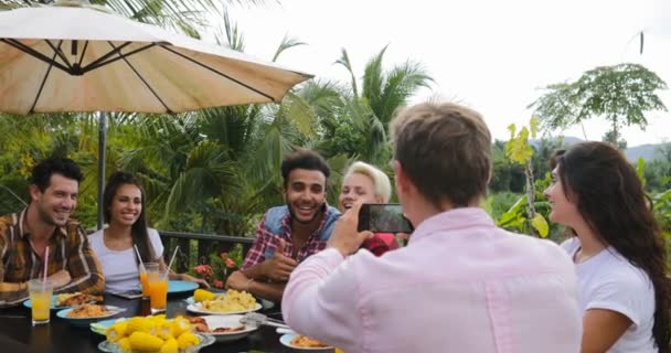 Homem tirando foto de casal no celular pessoas de telefone inteligente conversando sentado na mesa ao ar livre comendo no terraço grupo de jovens amigos feliz sorrindo — Vídeo de Stock