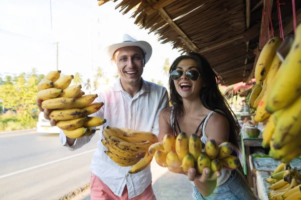 Joven pareja alegre sosteniendo racimo de plátanos en el mercado callejero felices turistas sonrientes en bazar de frutas asiáticas — Foto de Stock