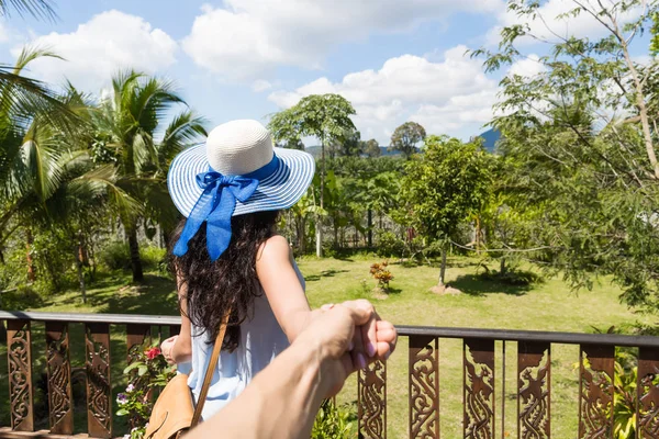 Volver Vista trasera de mujer joven en sombrero sobre hermoso paisaje tropical sosteniendo la mano del hombre — Foto de Stock