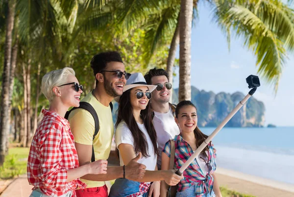 Gente grupo tomar selfie con acción cámara en palo mientras caminando en palmera parque en playa, feliz sonriente mezcla de raza amigos — Foto de Stock