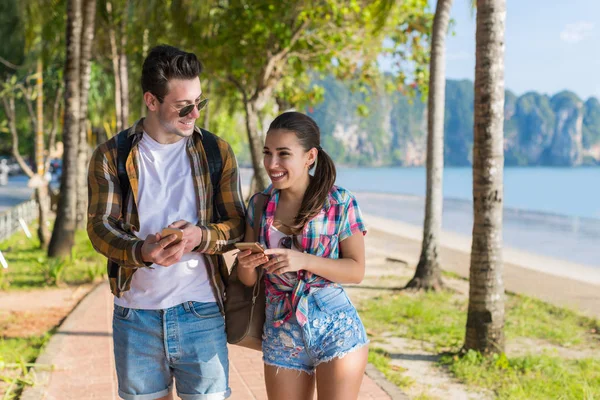 Випадкова пара Використовуйте мобільні смартфони у тропічному парку пальмових дерев, Щасливі усміхнені красиві молоді люди спілкуються онлайн на відкритому повітрі — стокове фото